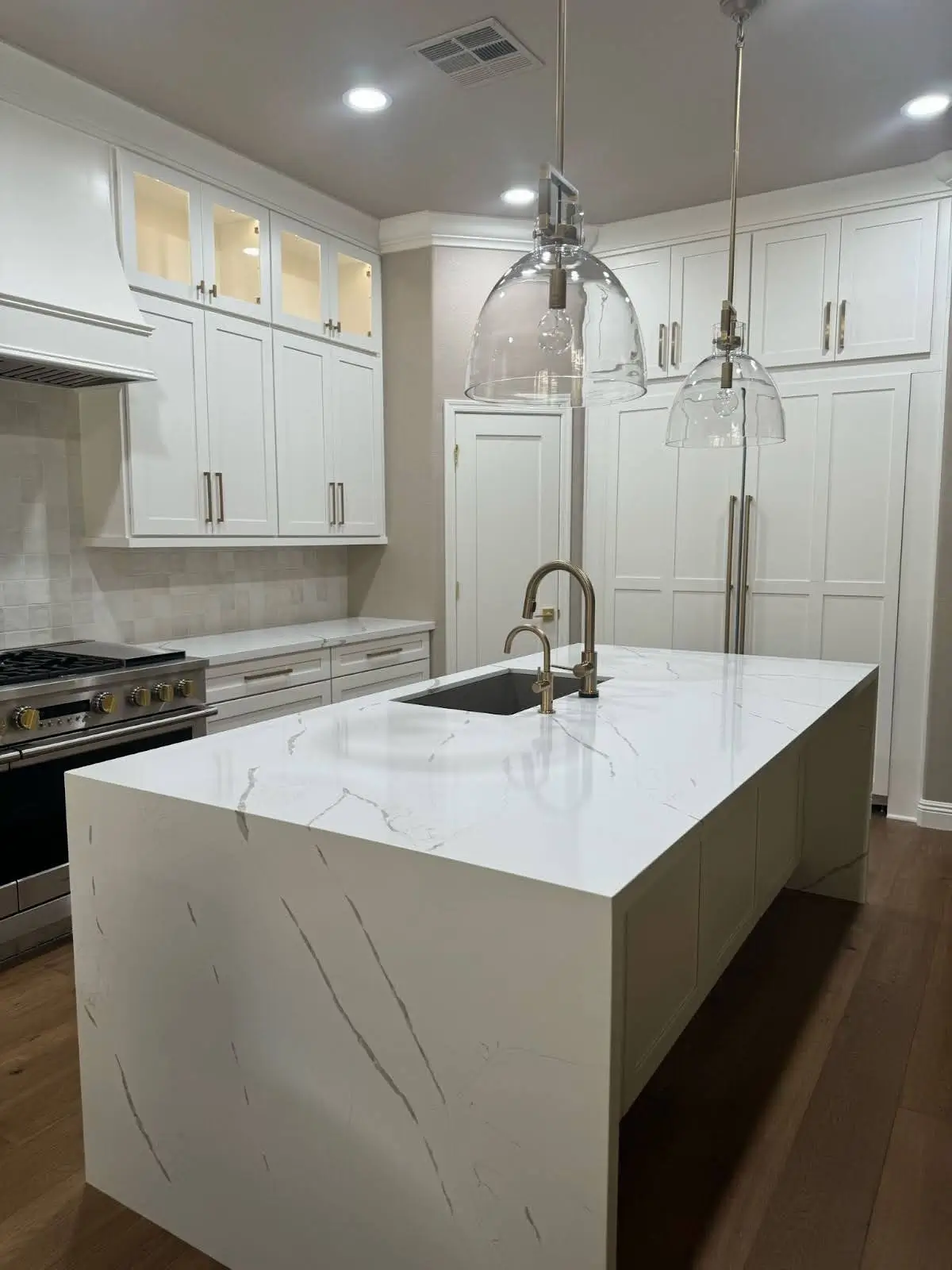 Elegant white kitchen with waterfall marble island, integrated sink, glass pendant lights, gold hardware, and seamless built-in cabinetry—blending appliances into a sleek, contemporary design.